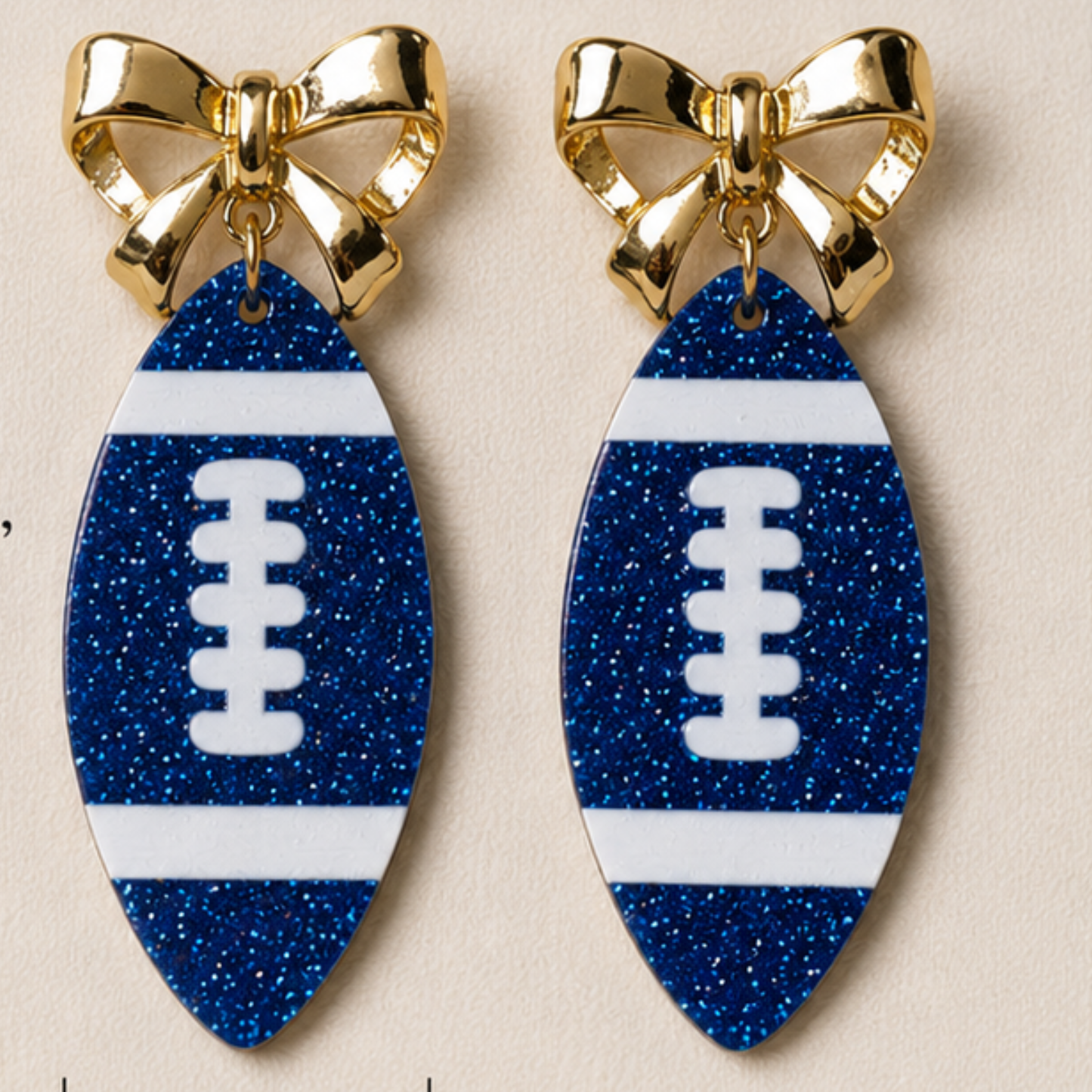 Pair of earrings with blue glittery football shapes and gold bows on a beige background soberafshop