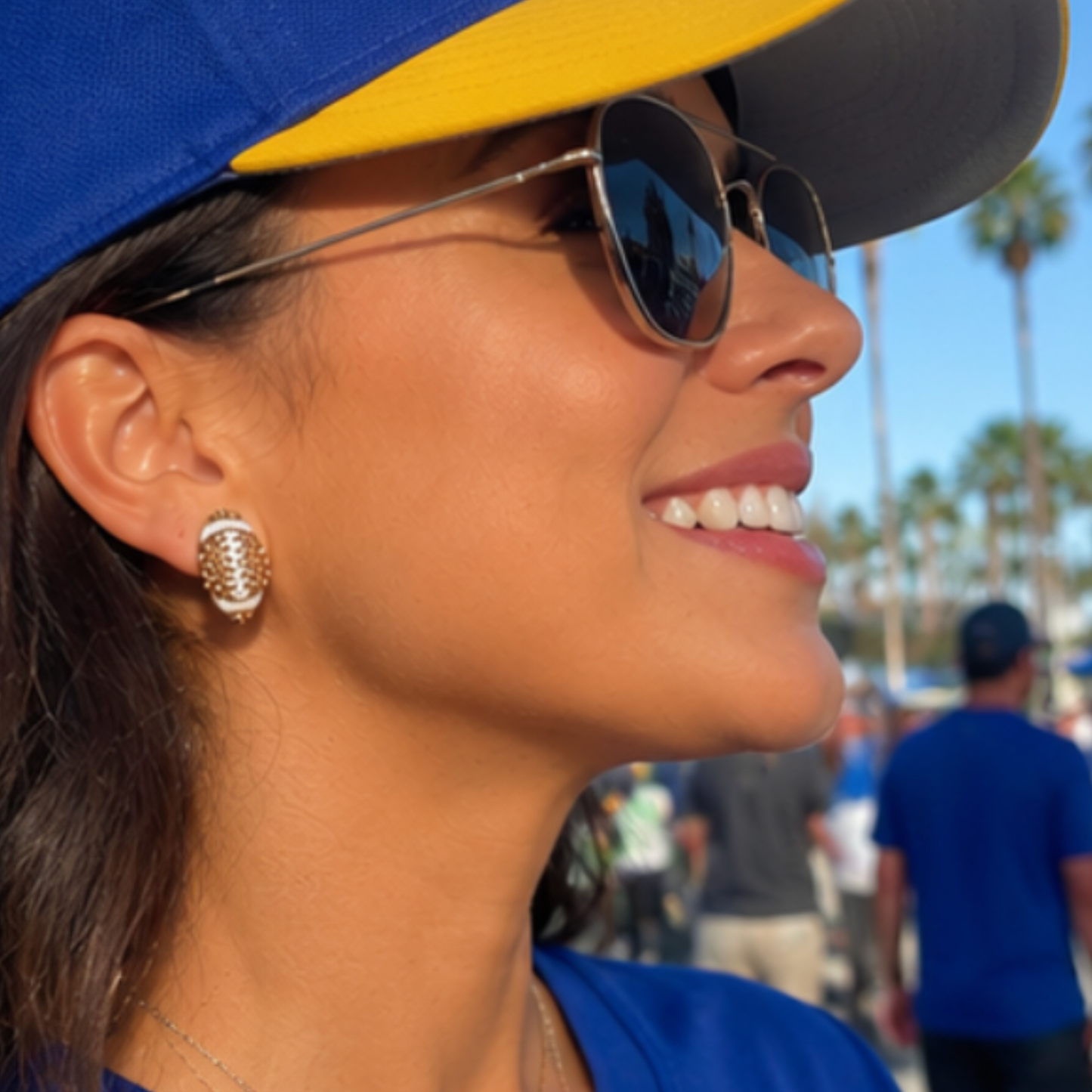 Woman wearing a blue and yellow cap and sunglasses with a blurred background of people and palm trees.wearing football rhinestone football stud earrings
\