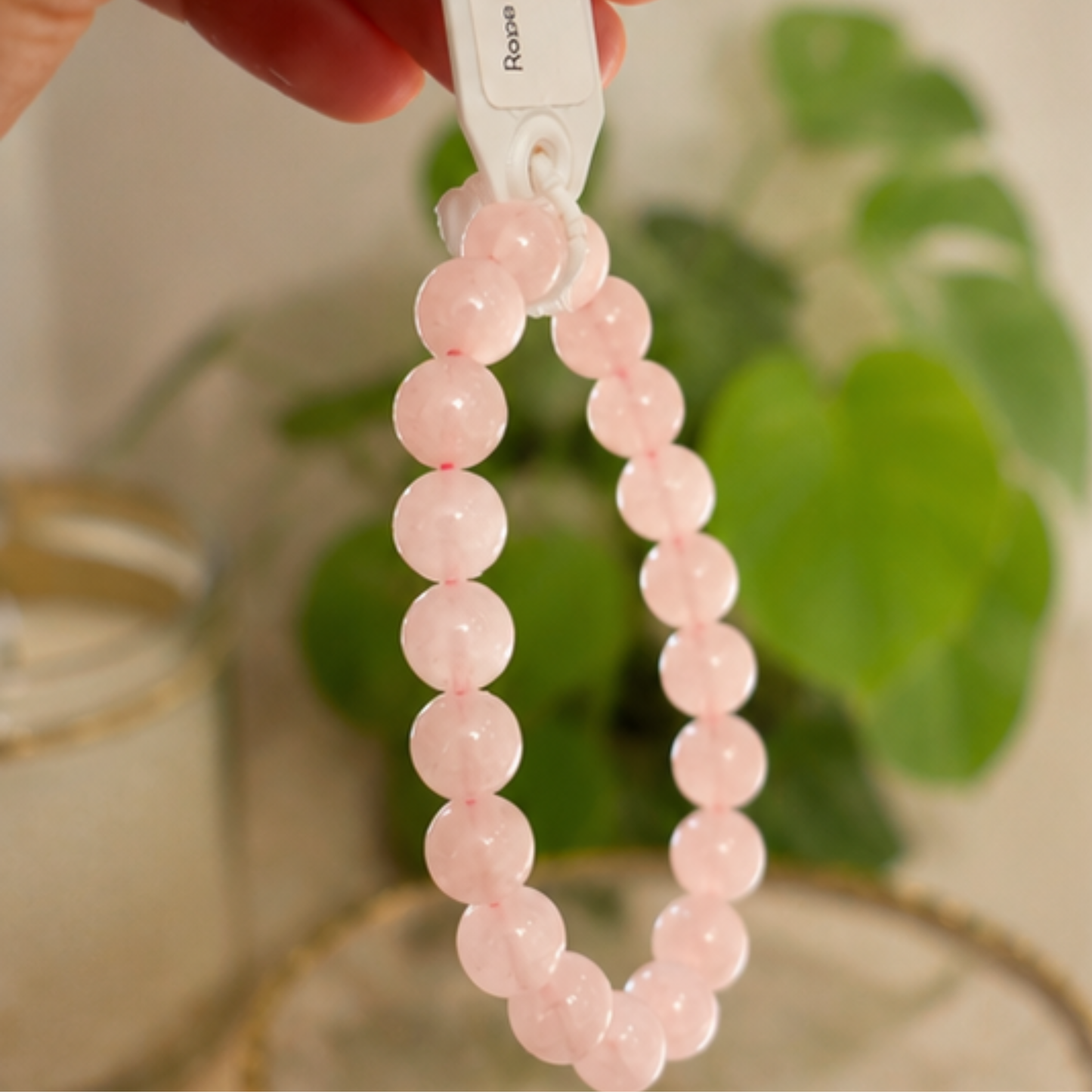 Pink rose quartz beaded bracelet  with a white tag held by a hand against a blurred green plant background